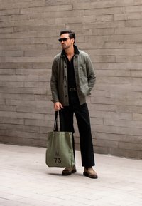 Olivgrüne Jacke über schwarzem Shirt, schwarze Hose, hält eine hellgrüne Einkaufstasche mit dem Aufdruck "M 73". Braune Slipper runden das Outfit ab.