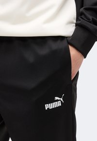 Χέρι ατόμου στην τσέπη μαύρου παντελονιού Puma, φοράει μαύρο και λευκό τοπ, εμφανίζοντας το λογότυπο Puma στο παντελόνι κοντά στο μηρό.