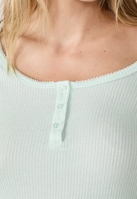 Jasno miętowy top z prążkowanej tkaniny, z okrągłym dekoltem wykończonym koronką oraz z listwą z trzema jasno niebieskimi guzikami.
