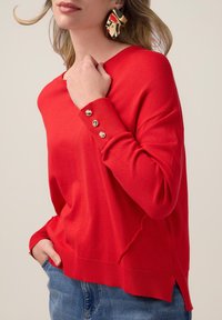 Maglione rosso lavorato a maglia con scollo rotondo, maniche lunghe e tre bottoni decorativi sul polsino. Abbinato a jeans blu con una fessura laterale.