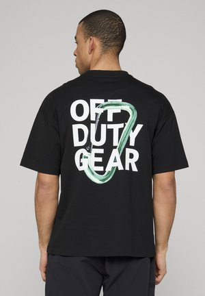Man met zijn rug naar de kijker, draagt een zwart T-shirt met witte tekst "OFF DUTY GEAR" en een grote groene karabijnhaak op de rug.