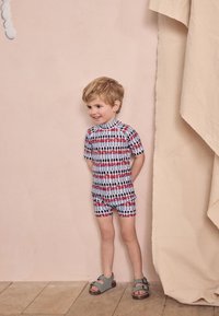 Kinder-Rashguard mit kurzen Ärmeln in Weiß mit bunten Spielzeugmustern, kombiniert mit passenden Shorts und grauen Sandalen auf Holzboden.