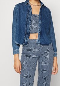 Camicia di denim blu scuro con chiusura a bottoni e orlo sfrangiato. Indossata sopra un top a righe cropped e pantaloni abbinati a vita alta.