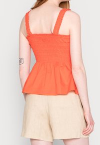 Top arancione smockato con spalline regolabili, orlo a peplum svasato e tessuto testurizzato; abbinato a pantaloni beige.