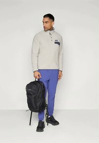 Ανοιχτό γκρι φλις πόλο με ναυτικό μπλε τσεπάκι στο στήθος, συνδυασμένο με μωβ παντελόνι. Μαύρο σακίδιο διαθέτει λογότυπο και ρυθμιζόμενους ιμάντες. Μαύρα αθλητικά παπούτσια.