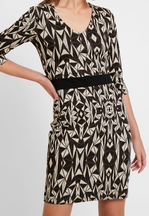 Kvinde iført en knælængde kjole med sort-hvide geometriske mønstre og en ensfarvet sort taljebånd, poserer med den ene hånd på hoften.