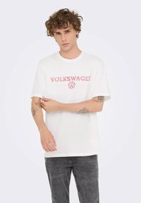 Joven con pelo rizado que lleva puesta una camiseta blanca de Volkswagen y unos pantalones vaqueros oscuros ajustando la manga, con tatuajes visibles en el brazo frente a un fondo sencillo.