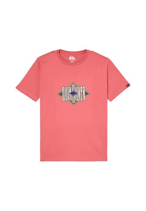 Chłopiec z kręconymi włosami stoi, ubrany w koralowy t-shirt z geometrycznym logo, luźne jasnoniebieskie dżinsy i czarne trampki z białymi podeszwami.