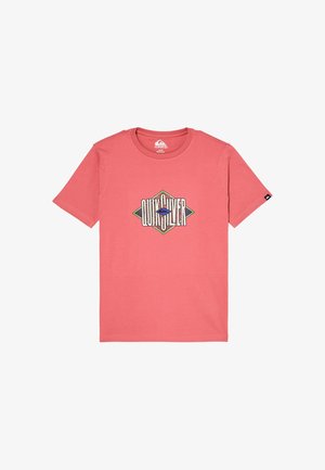 Chłopiec z kręconymi włosami stoi, ubrany w koralowy t-shirt z geometrycznym logo, luźne jasnoniebieskie dżinsy i czarne trampki z białymi podeszwami.