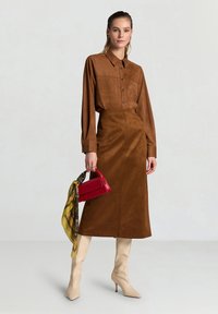 Brown suede shirt and midi skirt with a red handbag and a yellow patterned scarf. Beige high-heeled boots. Smooth texture, dapper design.