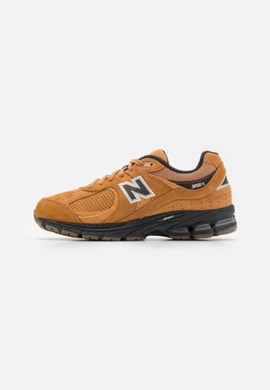 Bruine suède New Balance 2002R sneaker met zwarte gevoerde zool en wit logo aan de zijkant, getoond vanaf de buitenkant op een witte achtergrond.