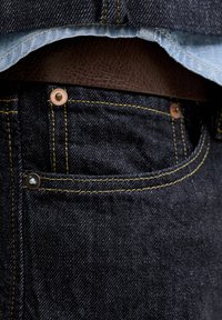 Καφέ δερμάτινη ζώνη περασμένη από τα θηλύκια σκούρου μπλε τζιν, ορατή μέση και ραφές μπροστινής τσέπης με μεταλλικά καρφιά.