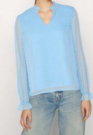 Jasnoniebieska przezroczysta bluzka w mały wzór kropek, z falbankowanym kołnierzem w kształcie litery V, długie rękawy z falbankowanymi mankietami, noszona z wyblakłymi niebieskimi dżinsami.