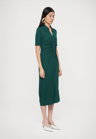 Zelené šaty na zapínanie s priliehavým živôtikom, našuchorenými detailmi, krátkymi rukávmi a dĺžkou ku kolenám. Nosí sa s bielymi slip-on topánkami.