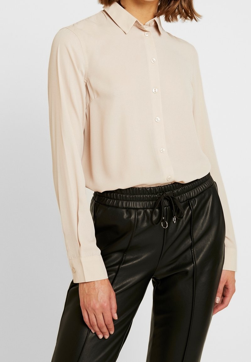 Personne portant une chemise beige à manches longues boutonnée, rentrée dans un pantalon en cuir noir à cordon, sur un fond uni.