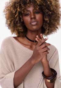 Beige gebreide trui met een losse pasvorm en opgerolde mouwen, voorzien van een zwarte choker ketting met een hart hanger en gelaagde armbanden.