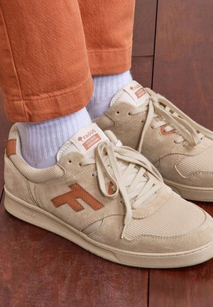 Zapatillas de gamuza beige con logo marrón y cordones blancos, usadas con calcetines blancos y pantalones naranjas, sobre un suelo de madera.