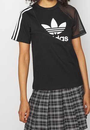 Femme portant un t-shirt Adidas noir avec des rayures blanches et des détails en mesh, assorti à une jupe plissée à carreaux noire et blanche.