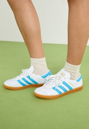 Fehér sportcipők világoskék csíkokkal, gumitalppal és fűzős dizájnnal. Arany logóval és sima bőrhatású felülettel rendelkezik.