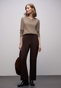 Beżowa prążkowana bluzka z długim rękawem i dekoltem w serek, w połączeniu z brązowymi, dopasowanymi spodniami i czarnymi botkami. Modelka trzyma ciemnobrązową torbę.