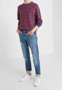 Gestreifter Pullover in Rot und Marineblau mit langen Ärmeln, kombiniert mit hellblauen, zerfetzten Denim-Jeans und weißen Sneakers. Lässiger Schnitt, modernes Design.