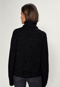 Mustat, paksut neuleet turtleneck-pusero, jossa on rento istuvuus, ja jossa on valahtavat olkapäät sekä teksturoitu kangas. Takaa näkyy ribbaus hihansuissa.