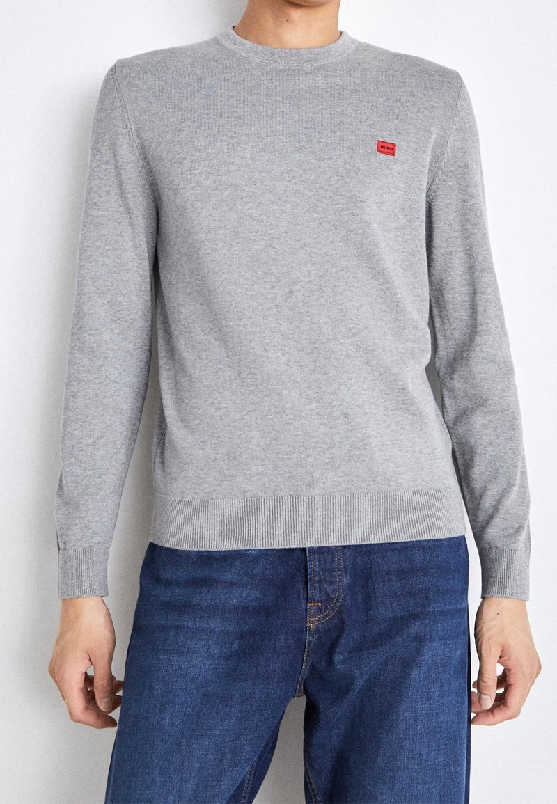 Szary sweter z długim rękawem wykonany z dzianiny, z okrągłym dekoltem i małą czerwoną naszywką z logo na lewej piersi. Mankiety i dół swetra wykończone ściągaczem.