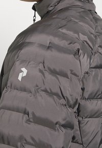 Giacca imbottita grigia con una texture liscia e lucida. Presenta un logo sulla spalla e un design con colletto alto per un maggiore calore.