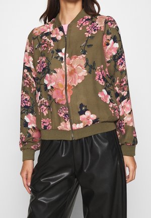 Kobieta w oliwkowej bomberce z różowo-czarnym kwiatowym wzorem i czarnych spodniach z sztucznej skóry, stojąca na jednolitym tle.