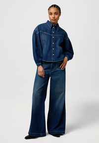 Veste en denim avec un col structuré, fermeture à boutons sur le devant et manches bouffantes, associée à un jean en denim à jambes larges avec poches arrière.