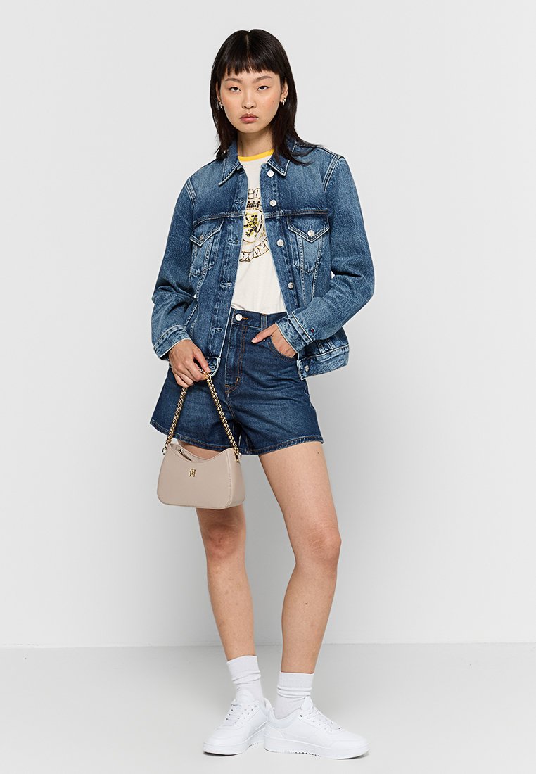 Jeansjacke mit heller Waschung, weißes Grafik-T-Shirt, dunkle Jeansshorts, weiße Sneaker und eine beigefarbene Schultertasche mit goldenen Akzenten.