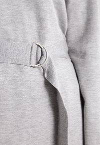 Un tessuto grigio con una texture morbida presenta un passante laterale che attraversa un anello in metallo argentato, creando un dettaglio strutturato nel design.