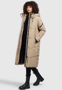 Beige langer Puffer-Mantel mit Kapuze, Reißverschluss-Taschen und Druckknöpfen. Schwarze Akzentdetails entlang des Reißverschlusses. Über einem schwarzen Oberteil und einer Hose getragen.