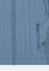 Giacca isolante blu chiaro con cuciture verticali, chiusura a zip completa e una tasca laterale con zip per un deposito sicuro. Texture liscia e lucida.