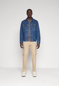 Denimjack in medium blauw, grijze T-shirt, beige cargobroek met zakken en witte sneakers. Staande tegen een neutrale achtergrond.