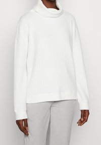 Weißer gerippter Rollkragenpullover mit langen Ärmeln, lockerer Passform, strukturierter Stoff und dezentes Strickmuster. Kombiniert mit hellgrauen Hosen.