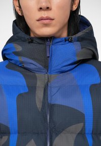 Jachetă căptușită cu un model abstract albastru și gri, cu guler înalt și glugă ajustabilă. Materialul are o finisare texturată.