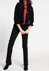 Schwarze Jeansjacke mit silbernen Knöpfen, kombiniert mit einem roten T-Shirt. Schwarze Schlagjeans und schwarze Knöchelschuhe runden das Outfit ab.