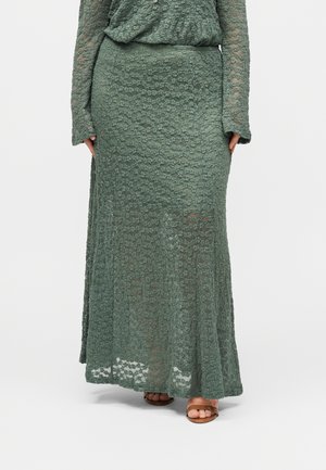 Langer grüner Spitzenrock, getragen mit passendem Spitzenoberteil, zeigt Füße in braunen Sandalen mit offenen Zehen auf weißem Hintergrund.