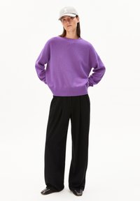 Frau mit Händen hinter dem Rücken, die eine hellgraue Mütze, einen lila Pullover, schwarze weit geschnittene Hosen und schwarze Schuhe mit Nieten trägt, auf weißem Hintergrund.