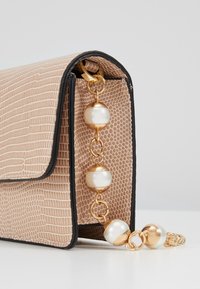 Malá béžová texturovaná kabelka s černými detaily a ozdobným zlatým řetízkem s velkými bílými a zlatými korálky.