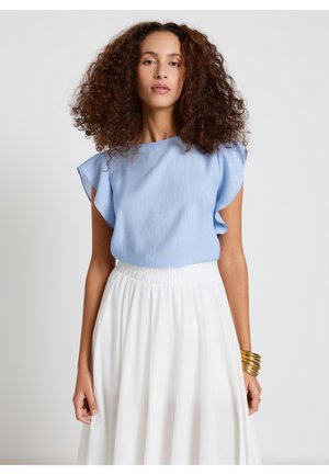 Frau mit lockigem Haar, die eine hellblaue ärmellose Bluse und einen hoch taillierten weißen Rock trägt, mit goldenen Armreifen am Handgelenk.