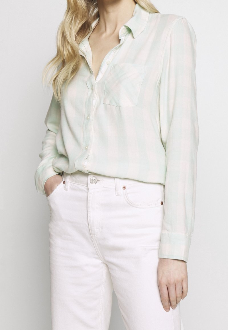 Camisa de cuadros verde claro y blanca con un bolsillo en el pecho, hecha de una tela suave, combinada con pantalones blancos.