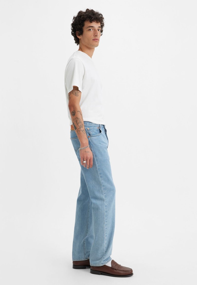 Helle blaue Denim-Jeans mit einem entspannten Schnitt, ausgestattet mit einem Fünf-Taschen-Design und subtilen Nähten. Kombiniert mit einem schlichten weißen T-Shirt und braunen Mokassins.