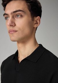 Sort polo t-shirt med en kort, V-formet krave. Blødt, struktureret stof. Tæt fit, der fremhæver halsudskæringen og hagen. Neutral baggrund.