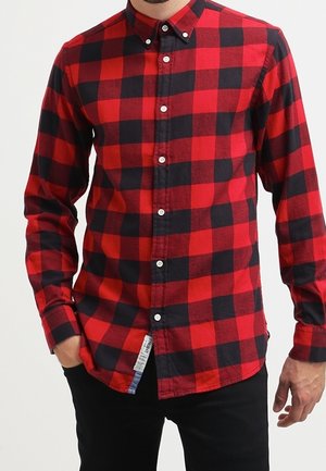 Homme portant une chemise à manches longues à carreaux rouges et noirs avec des boutons blancs, associée à un pantalon noir, debout avec une main dans la poche.