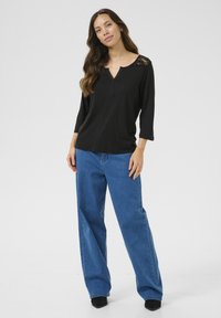 Haut noir à manches longues avec accent en dentelle, décolleté en V et patte de boutonnage, associé à un jean en denim bleu ample et des chaussures noires.