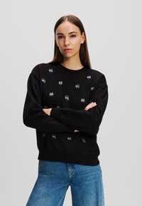 Schwarzer Baumwollpullover mit Rundhalsausschnitt und langen Ärmeln, verziert mit kleinen gestickten Gesichtsmotiven in Weiß und Grau.