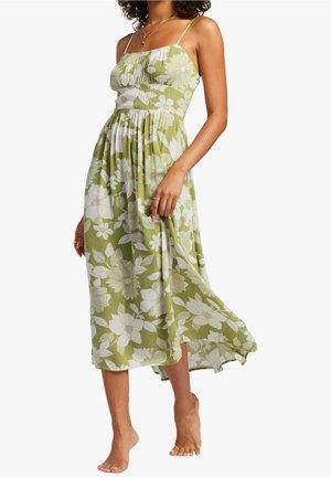 Robe maxi florale verte avec bretelles spaghetti ajustables, corsage froncé et ourlet fluide ornés de motifs de fleurs blanches. Modèle pieds nus.