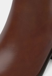 Botină de piele maro, la gleznă, cu o textură netedă, o siluetă curbată și o talpă din cauciuc negru. Prezintă o cusătură minimă și nu are hardware vizibil.